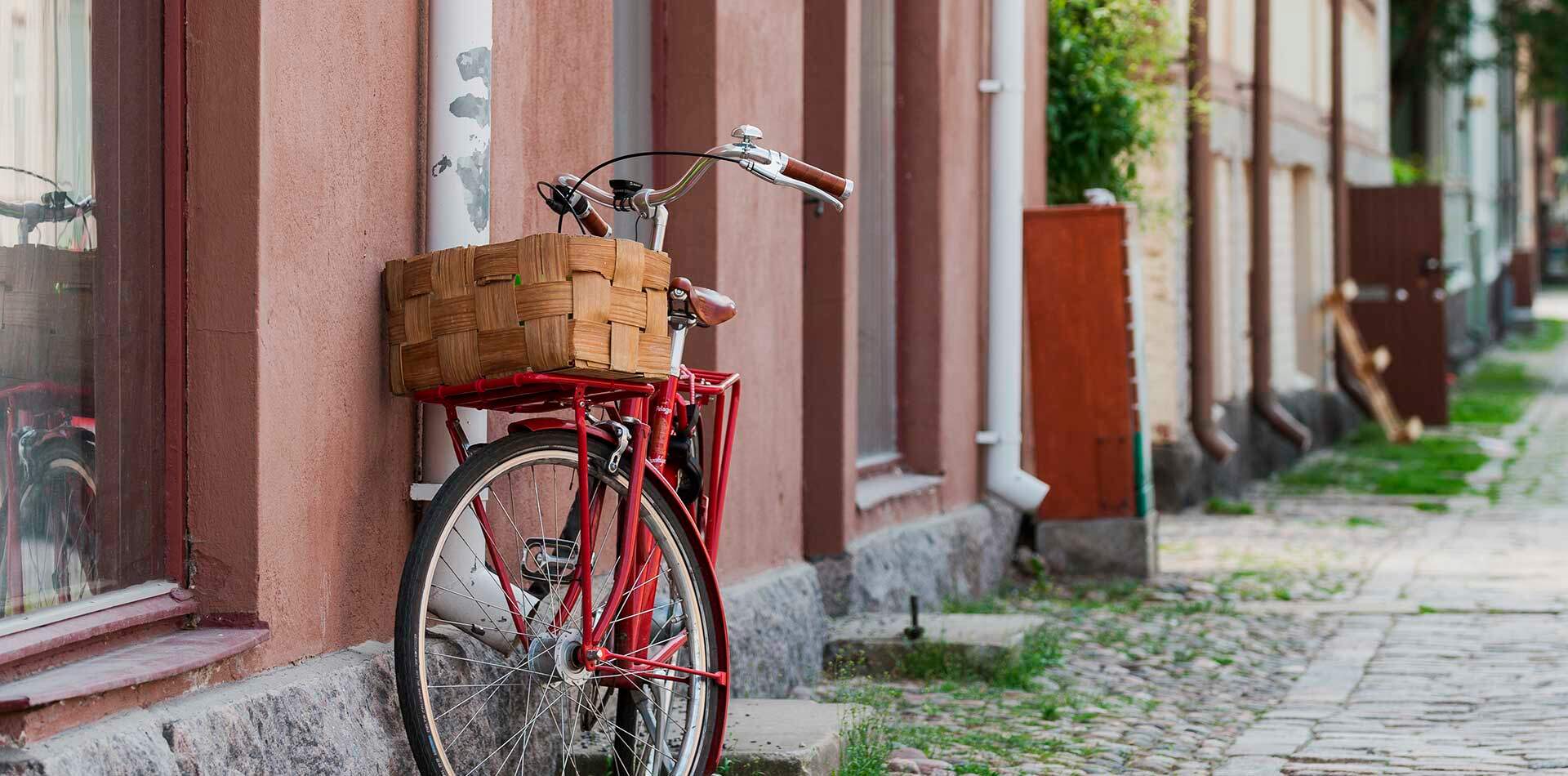 Valokuva - Punainen polkupyörä, jossa on korillinen etuosa, nojaa punatiiliseen seinään. Taustalla näkyy mukulakivikatu ja rakennuksia.