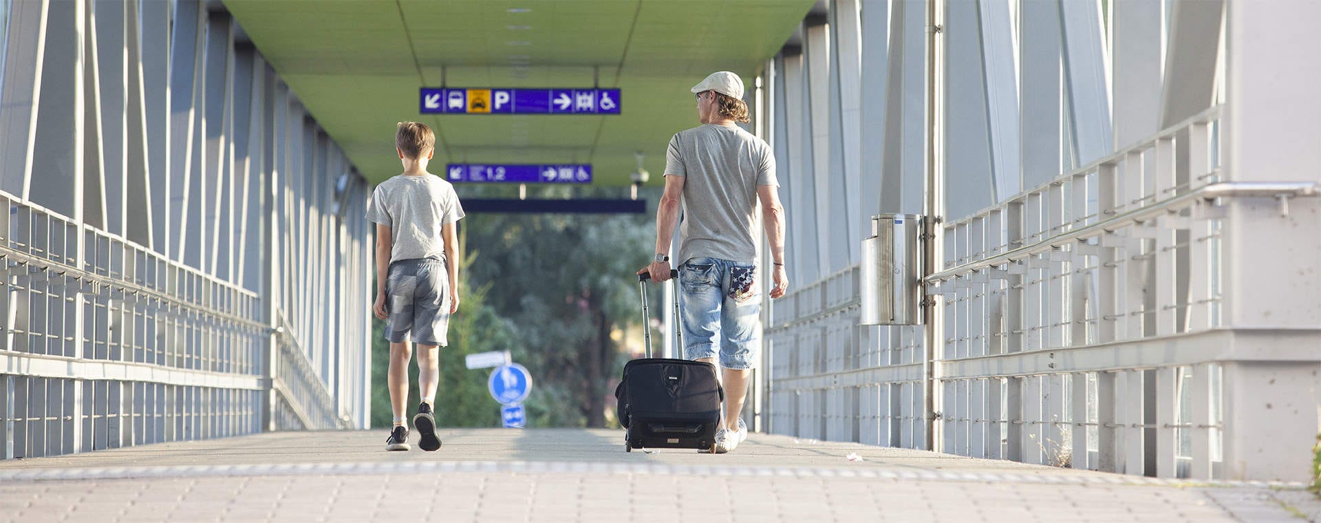 Mies ja poika kävelevät modernilla ylikulkusillalla Leinelän juna-asemalla. Mies vetää mustaa matkalaukkua, ja yläpuolella näkyvät opasteet ohjaavat eri suuntiin. Taustalla näkyy vehreää maisemaa ja aurinkoinen sää.