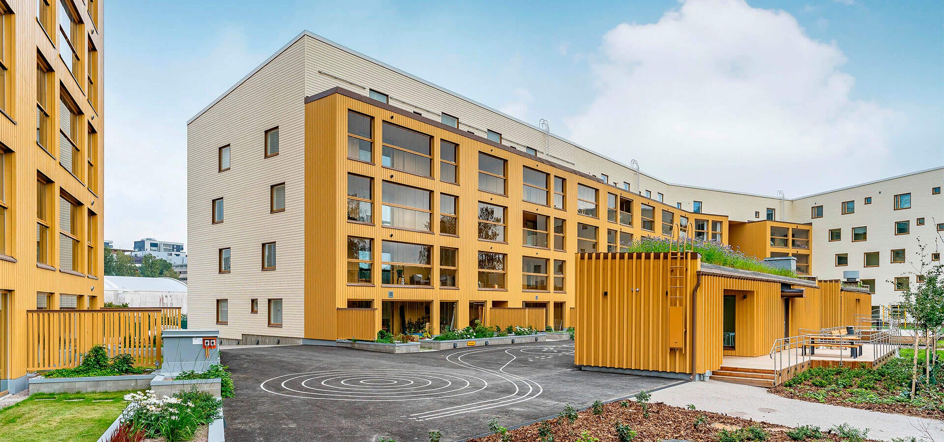 Valokuva Espoon Tuulikello 2:sta sisäpihan puolelta Vaaleankeltaiset kerrostalot vehreällä sisäpihalla, leikkipiirroksin kuvioitu asfalttipiha ja viherkattoinen piharakennus.