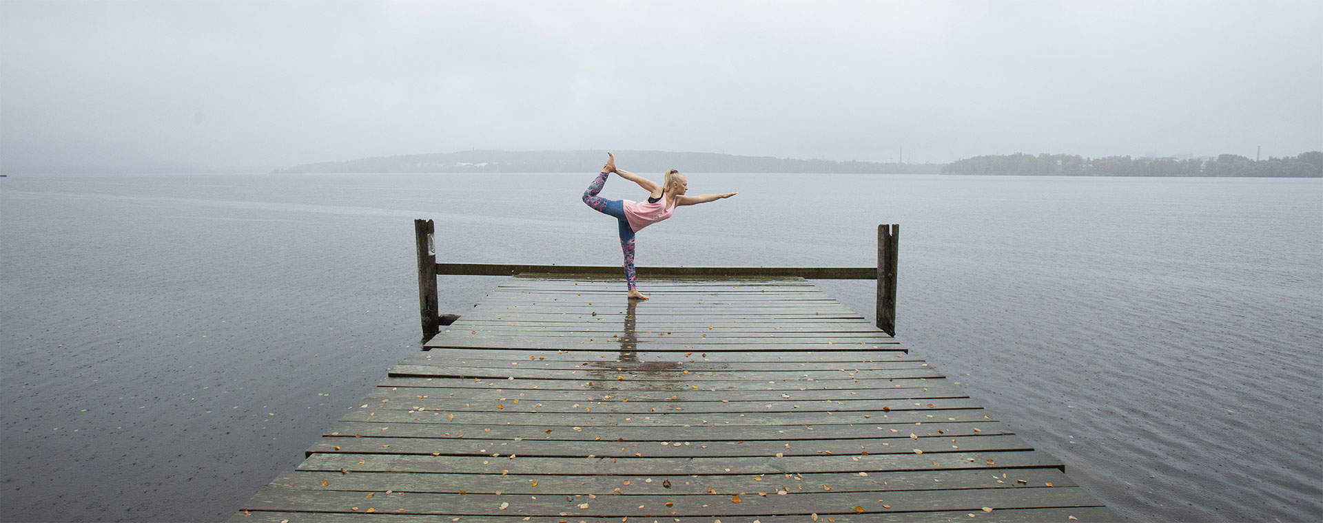 Kuva Härmälänrannasta. Joogaa harjoitteleva henkilö seisoo tasapainoasennossa märällä laiturilla harmaan järvimaiseman edessä. Taustalla näkyy sumuinen järvimaisema ja kaukaisia saaria.
