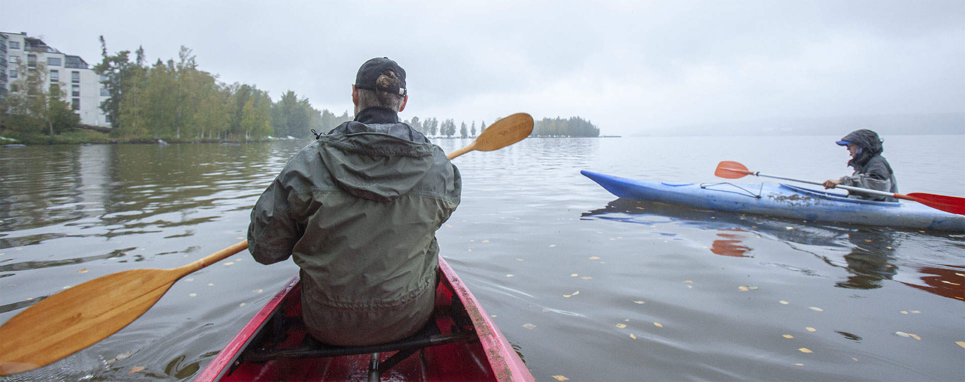 Henkilö meloo punaisessa kajakissa rauhallisella Pyhäjärvellä syksyisenä, sumuisena päivänä. Taustalla näkyy toinen sininen kajakki, rantapuita ja pilvinen taivas.