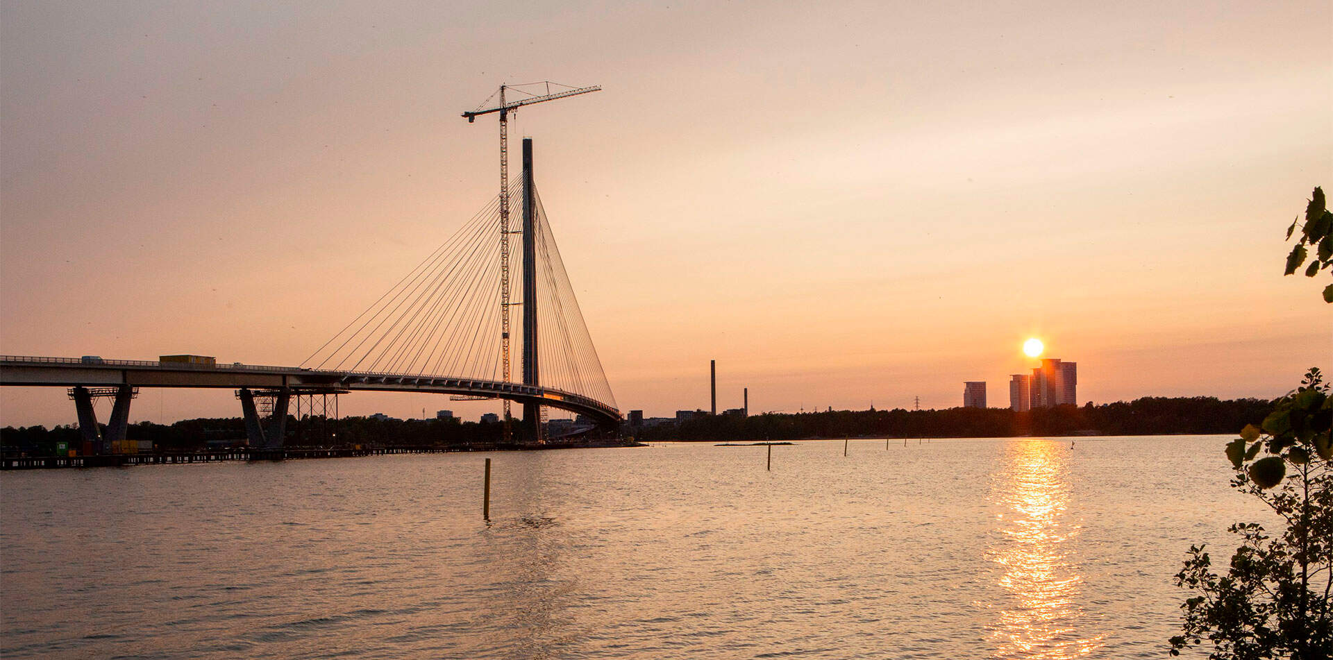 Auringonlasku Kruunuvuorensillan takana, sillan ylle kohoaa nosturi ja meren pinnassa heijastuu lämpimän oranssi valo. Taustalla siintävät korkeat asuintornit ja horisontin siluetti.