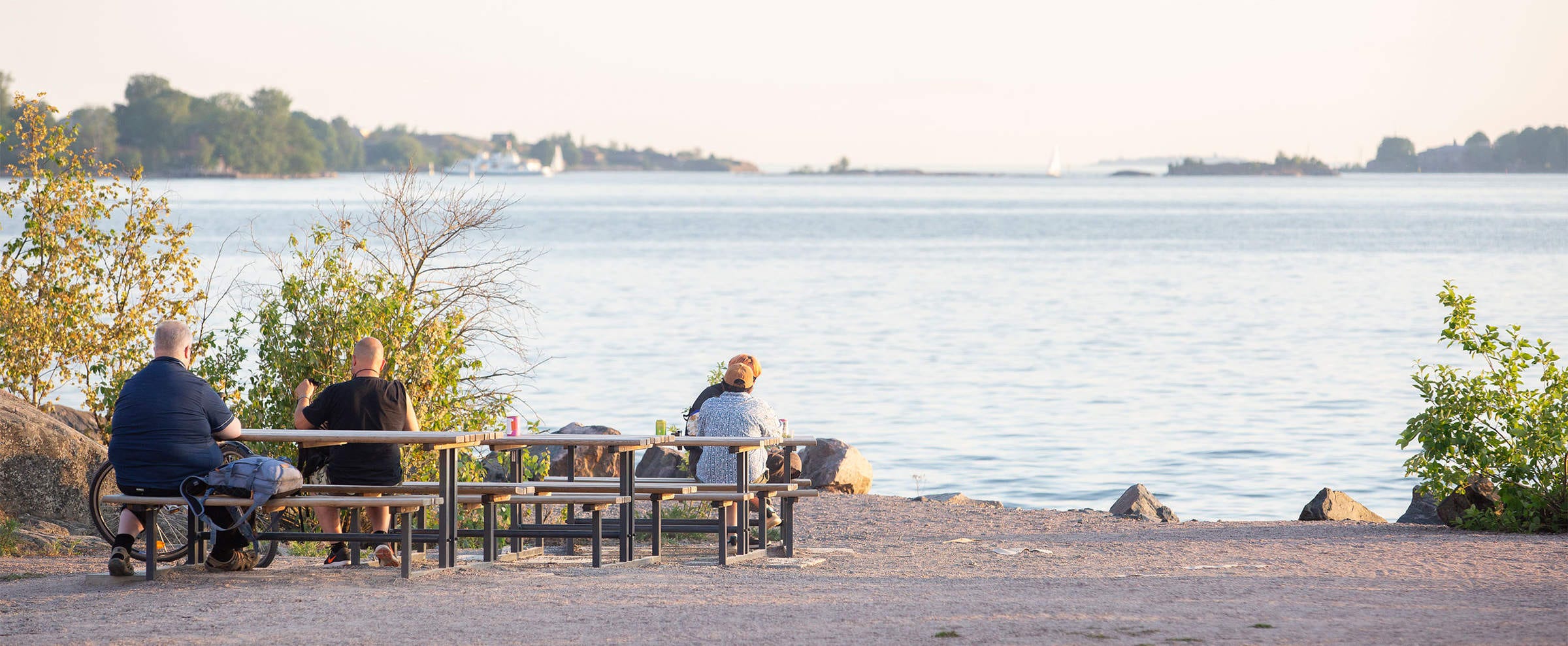 Iltapäivän aurinko valaisee Kruunuvuorenrannan rantamaiseman, jossa ihmiset istuvat penkillä nauttien kauniista merinäkymästä. Taustalla näkyy pieniä saaria ja purjeveneitä rauhallisella merellä.