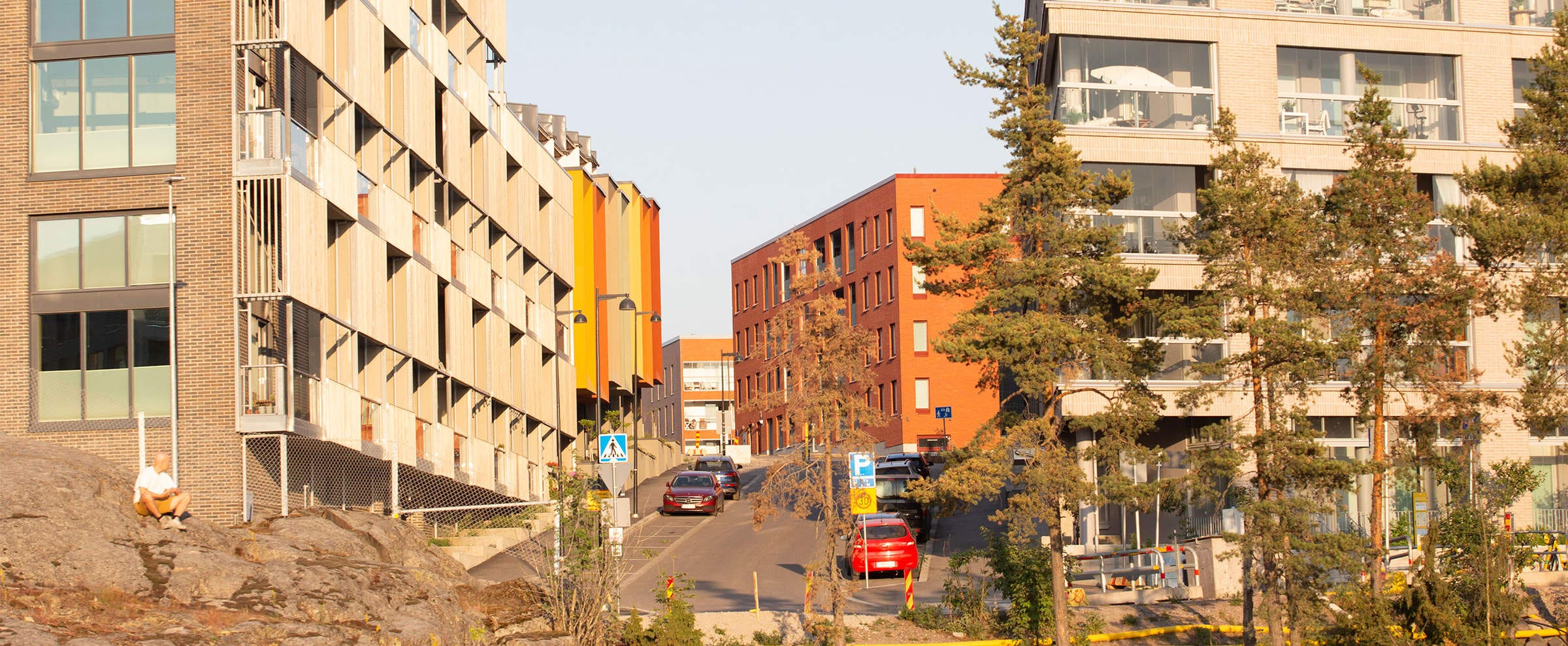 Katu Kruunuvuorenrannassa, jota reunustavat modernit kerrostalot. Rakennukset ovat värikkäitä ja oikealla lasiset parvekkeet erottuvat selkeästi. Kadun päässä näkyy punatiilinen rakennus. Metsäinen ympäristö kehystää kaupunkimaisemaa.
