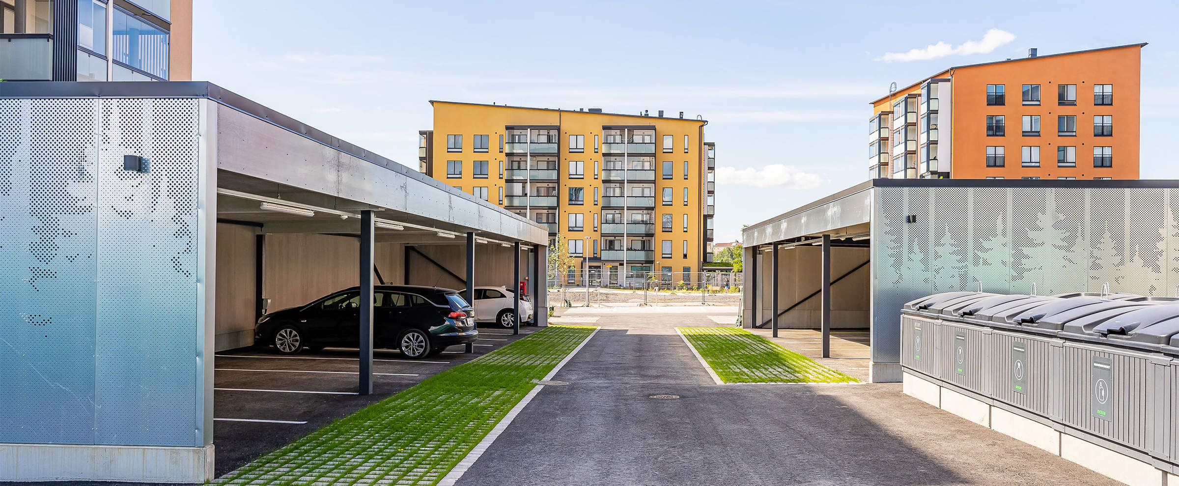 Moderni pysäköintialue Kirstinpuistossa kerrostalojen edustalla. Kuvassa näkyy katettuja autopaikkoja ja kierrätyspiste. Taustalla kohoavat keltaoranssit kerrostalot sinistä taivasta vasten.
