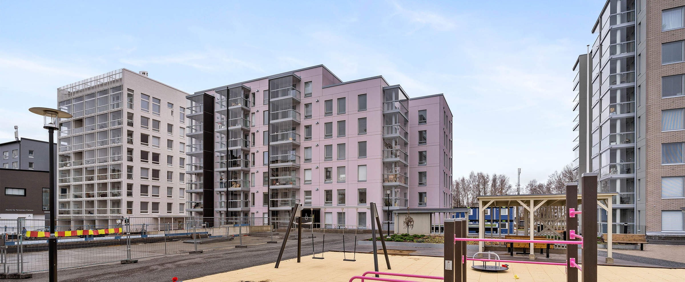 Uudisrakennuskohteen pihapiiri Härmälänrannassa, jossa on vaaleanpunaisia ja valkoisia kerrostaloja. Etualalla on moderni leikkipaikka, jossa on keinuja ja kiipeilytelineitä.