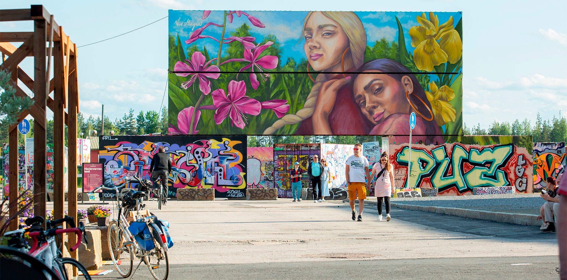 Värikäs graffitipuisto Hiedanrannassa, jossa suuri kukka-aiheinen muraali ja kävelijöitä aurinkoisena päivänä.