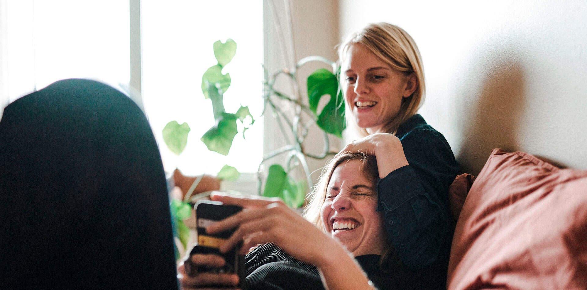 Tampereen Terra - uudiskohde Hiedanrannassa Valokuva - Kaksi ihmistä istuu sohvalla sisätiloissa, yksi henkilö käyttää matkapuhelinta. Taustalla on viherkasvi ikkunan edessä.