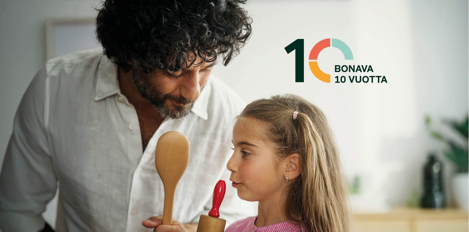 Valokuva - Aikuinen ja lapsi seisovat olohuoneessa. Aikuisella on kädessä puulusikka ja lapsella kaulin. Taustalla ruskea sohva ja taulu.