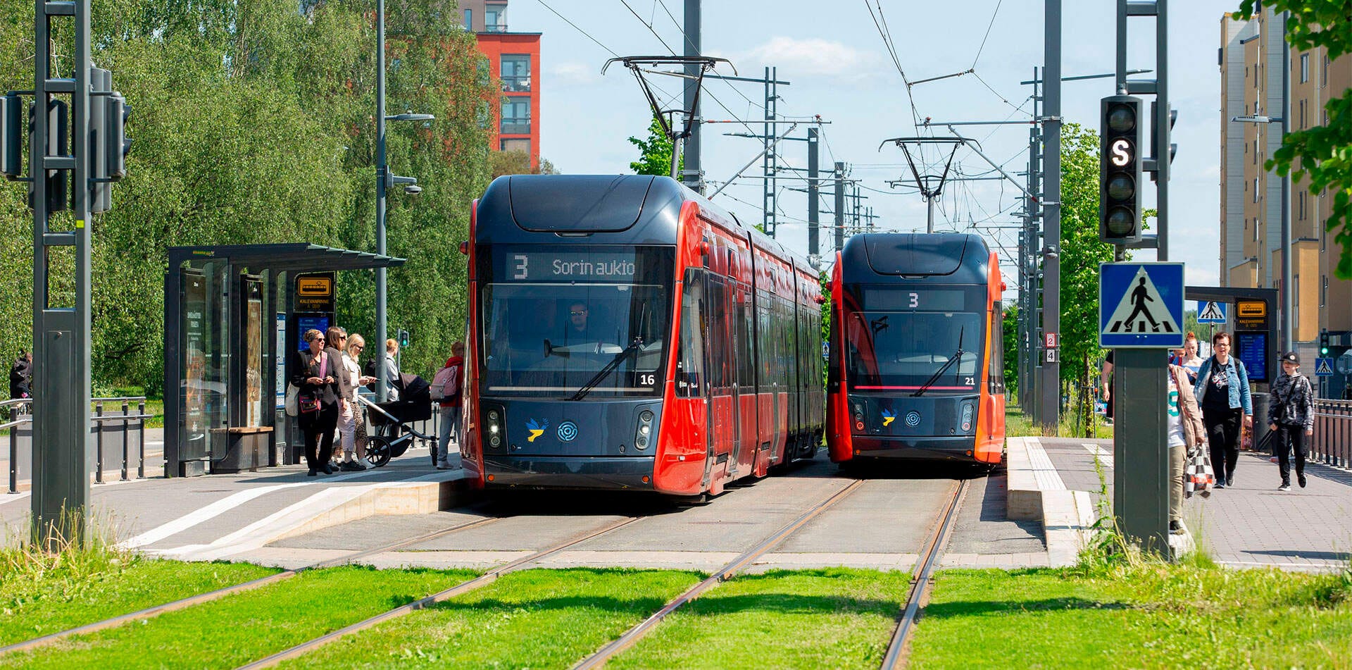 Kaksi punaista raitiovaunua pysähtyneenä Kalevanrinteen pysäkillä, matkustajia nousee kyytiin.